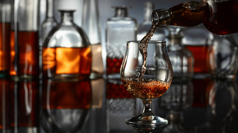 Brandy being poured from a bottle into a glass with decanters of brandy in the background.