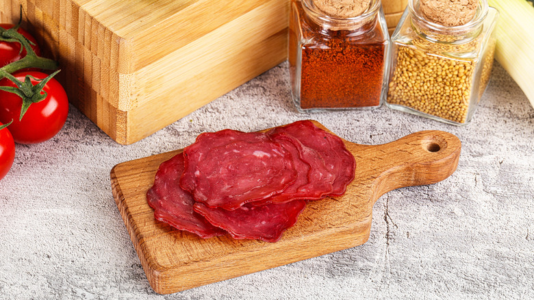 Pieces of pastrami on a wooden serving board next to spices and tomatoes