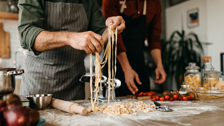 One person uses their hands to make pasta from scratch on a kitchen surface while another stands nearby