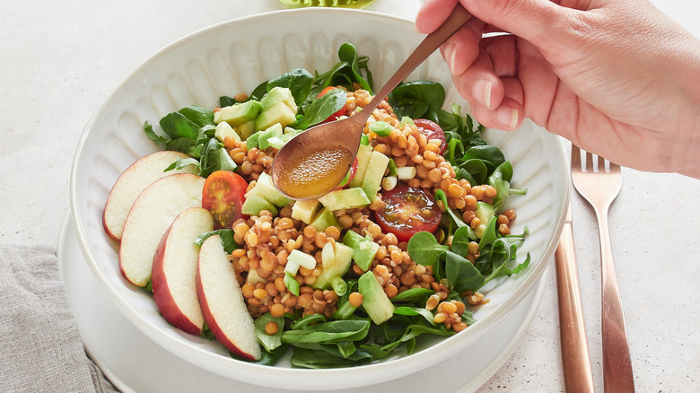 Someone adding vinaigrette to a salad containing apples, avocado, and tomatoes