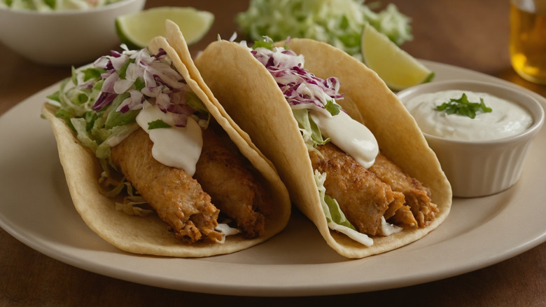 Two fried fish tacos on a white plate with a side of crema