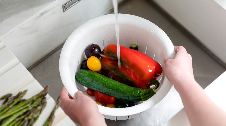 a woman washing a variety of vegetables in a white colander in her sink