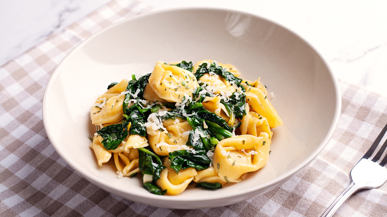 A bowl of tortellini with spinach and paremesan on a checked tablecloth