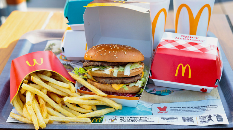 A McDonald's hamburger in the foreground with a packet of fries and McDonald's cups in the background