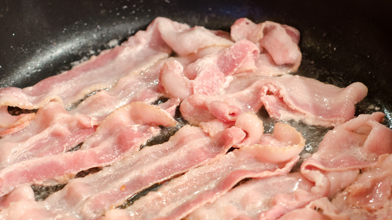 Strips of raw bacon are cooking in a pan