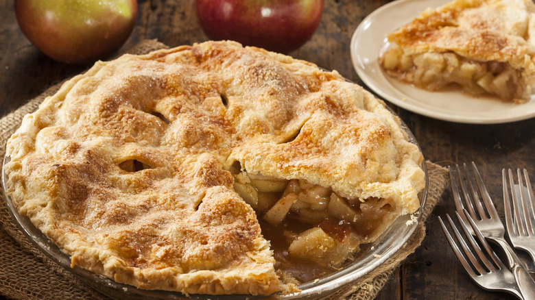 An apple pie with two apples behind it and a slice of apple pie beside it