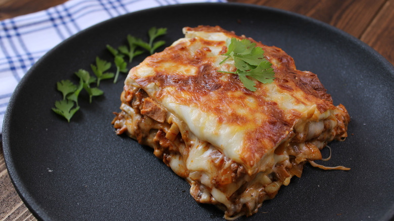 A slice of lasagna on a black plate with herbs on top