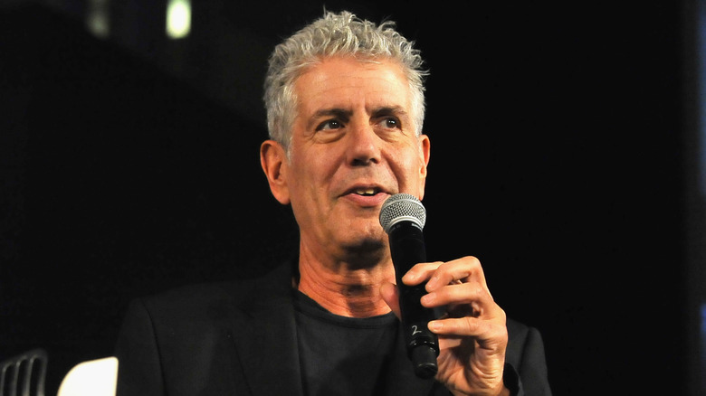 Anthony Bourdain holding a microphone and speaking at an event in a black blazer.