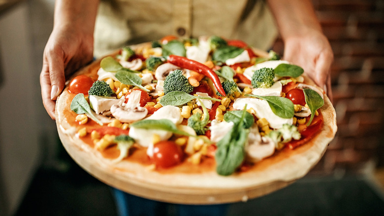 A person holding a pizza topped with green vegetables and mushrooms.