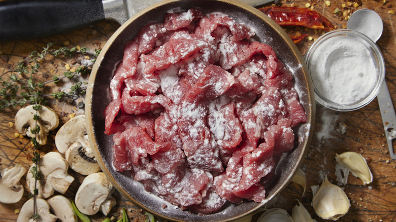 A bowl of raw steak tips sprinkled with baking soda sits on a wooden counter garnished with herbs, mushrooms, garlic, and a jar of baking soda.