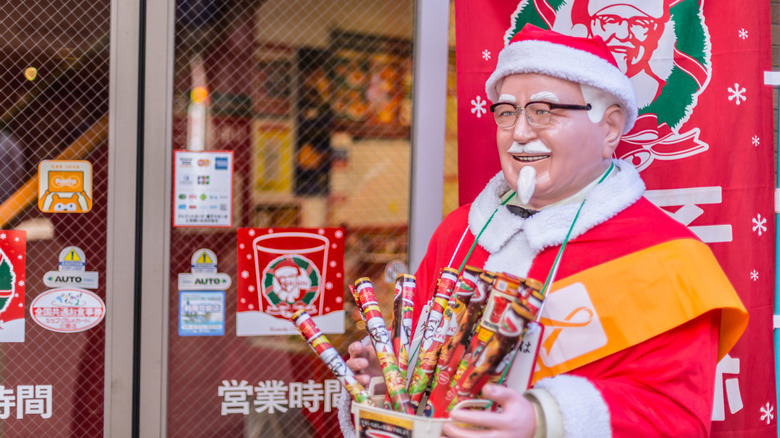 A statue of Colonel Sanders dressed as Santa Claus, holding KFC-branded treats in front of a KFC Christmas ad display