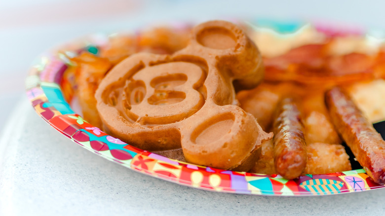 Mickey Mouse breakfast waffles with sausage and bacon on a colorful plate.