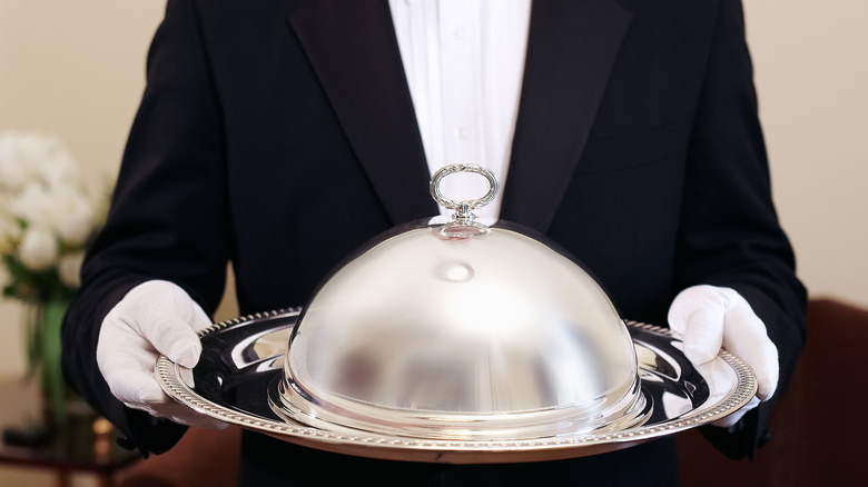 A server holding a silver cloche and tray bearing food.