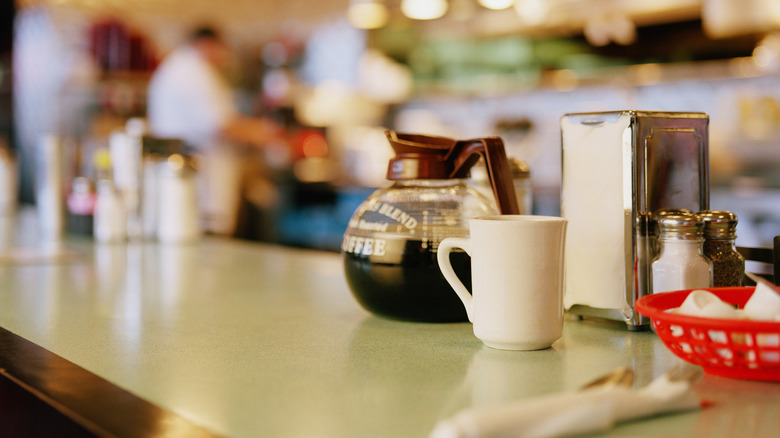 A counter inside a diner with a mug, a pot of coffee, a napkin dispenser, and salt and pepper shakers