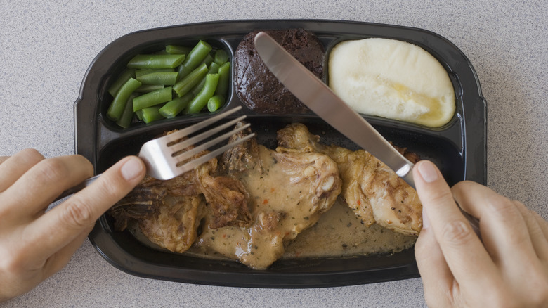 hands holding fork and knife above microwaved dinner in plastic tray