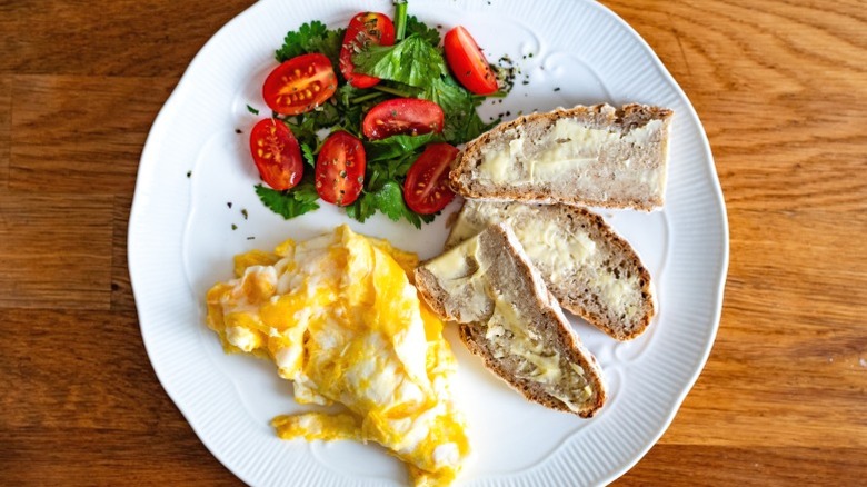 buttered bread, eggs, greens, and tomatoes on a white plate set on a wooden table