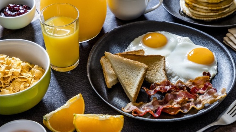 Breakfast plate with sunny-side up eggs, toast triangles, bacon, and a bowl of corn flakes with a glass of orange juice