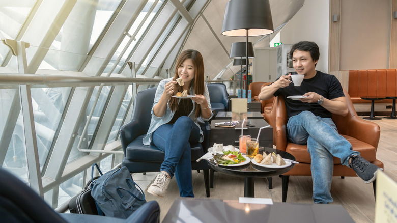 A man and a woman eat and drink while seated in an airport waiting area