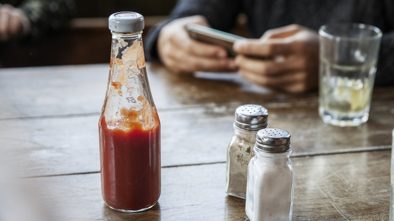 A glass bottle of ketchup on a table with salt and pepper shakers