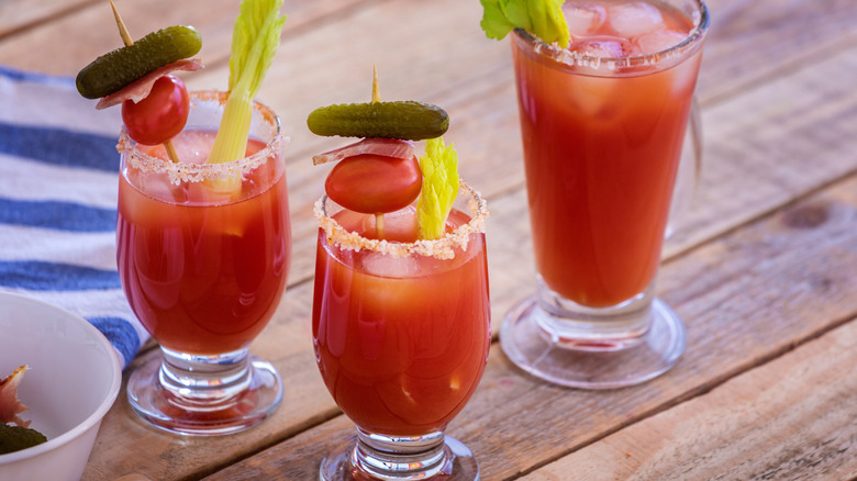 Three Bloody Mary cocktails on a wooden surface garnished with tomato, pickle, and celery