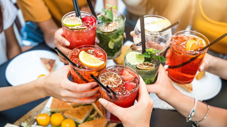 Seven hands clinking drinks around a table.