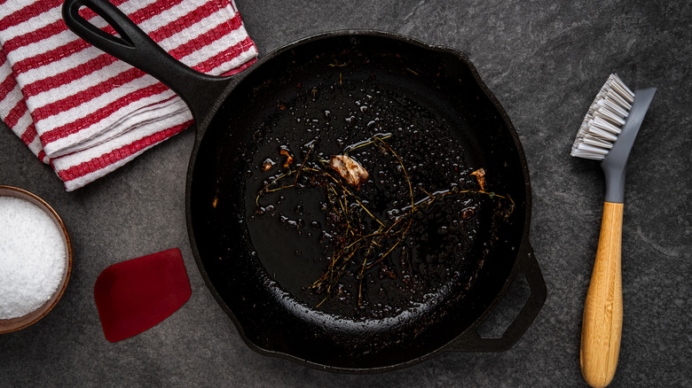 Dirty cast iron pan alongside brush, towel, and bowl of salt