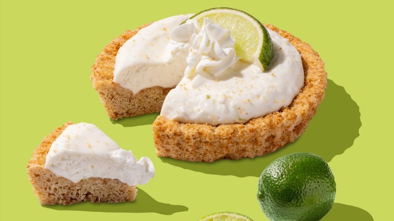 The Crumbl Key Lime Pie with a slice cut out of it next to a lime on a green background.