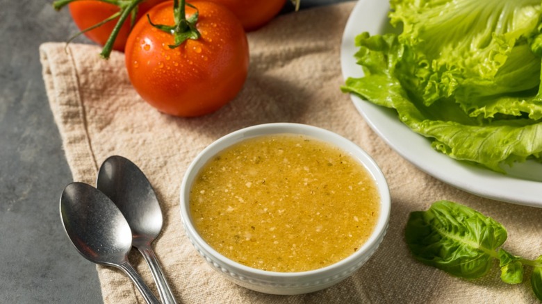 A white bowl of salad dressing rests on a countertop near a plate of lettuce, spoons, and tomatoes