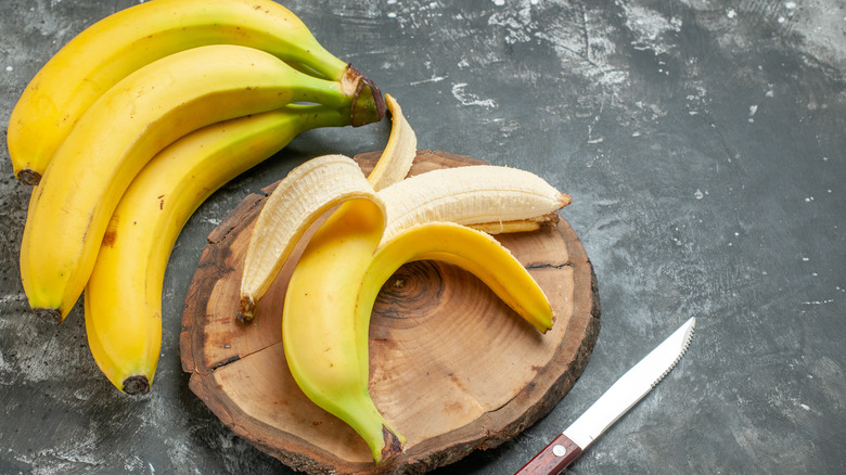 A peeled banana on a wooden board next to a bunch of bananas