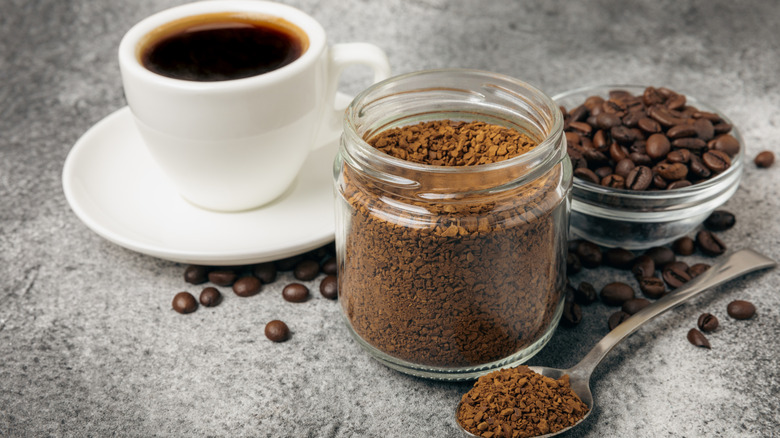 A jar of instant coffee with an espresso and a bowl of coffee beans in the background