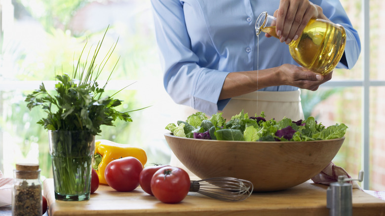 A person pours oil on a bowl of greens resting on a table with herbs and vegetables