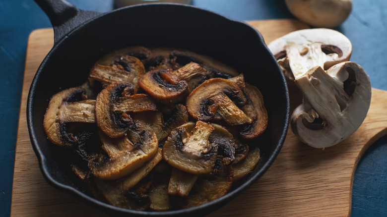 browned, sauteed mushrooms in a small frying pan beside raw sliced mushrooms over wooden cutting board