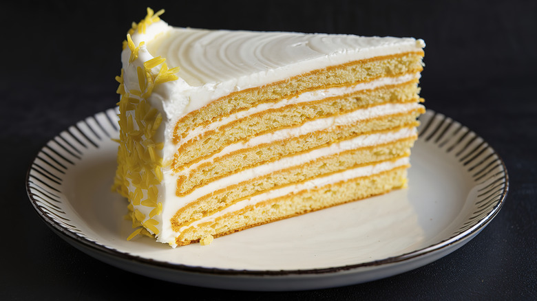 A slice of lemon layer cake sits on a white plate with black stripes around the edge, with a black background