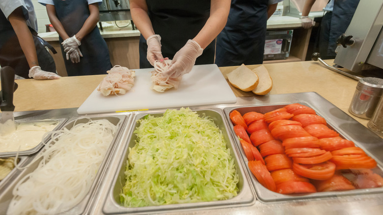 A Jersey Mike's worker assembles a sandwich in front of bins with sliced onions, lettuce, and tomatoes