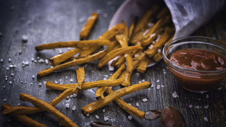 Thin sweet potato fries next to a glass bowl of ketchup on a black wooden surface.