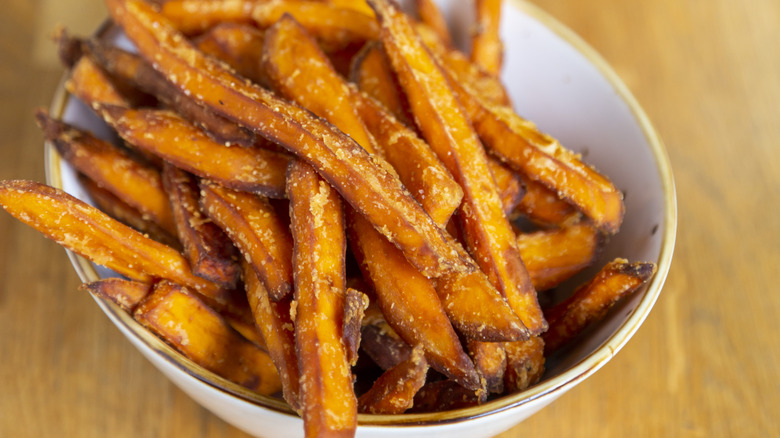 A close-up of a white bowl full of thin, crispy sweet potato fries on a wooden surface.