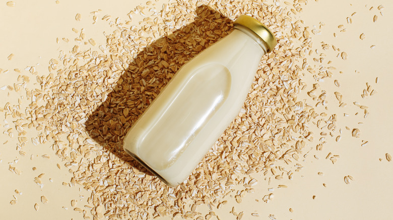 A glass bottle of oat milk and oatmeal flakes on a beige background.