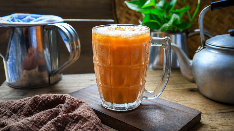 A mug of Thai iced tea rests on a table next to a teapot, strainer, and cloth