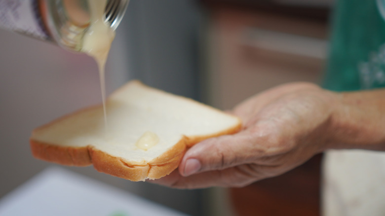 A person's hand holds a slice of bread as condensed milk is poured onto it