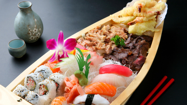 A sushi boat full of different foods with a sake cup and vessel nearby.