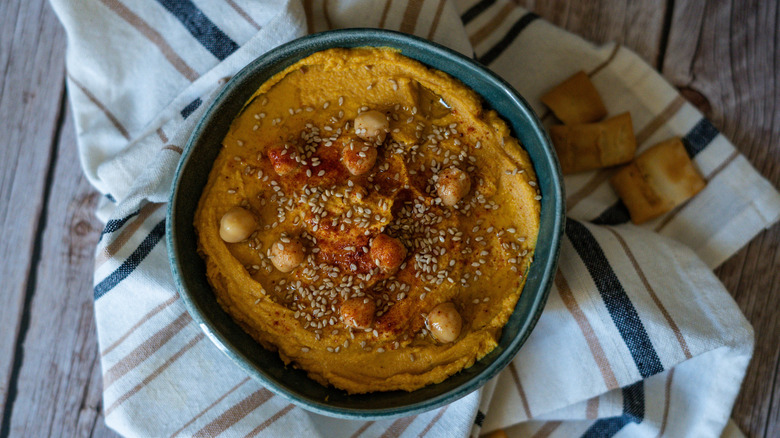 Dark bowl of pumpkin hummus with chickpeas and sesame seeds on striped cloth